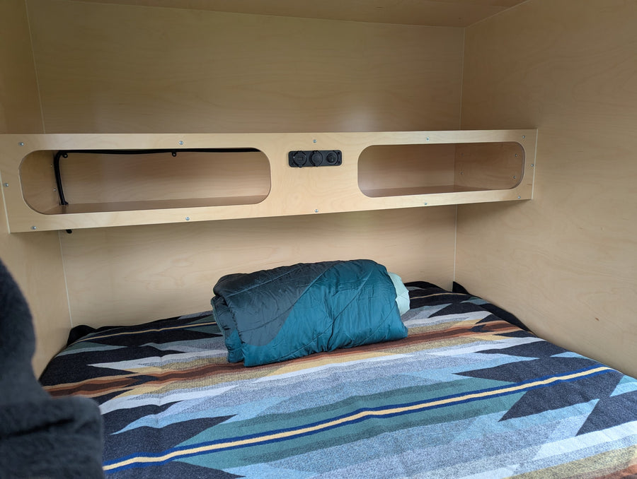 Cozy rv rental interior featuring a wooden shelf with outlets, cubbies, and a comfortable bed with a patterned blanket.