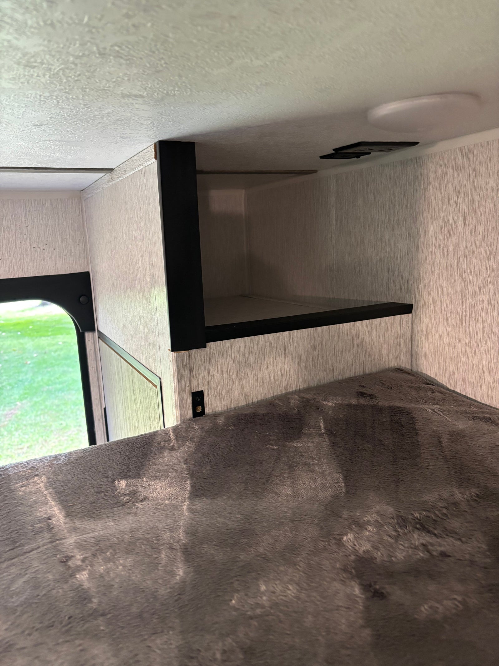 RV rental interior showing a cozy bunk bed with grey, textured bedding and an open black overhead storage shelf.