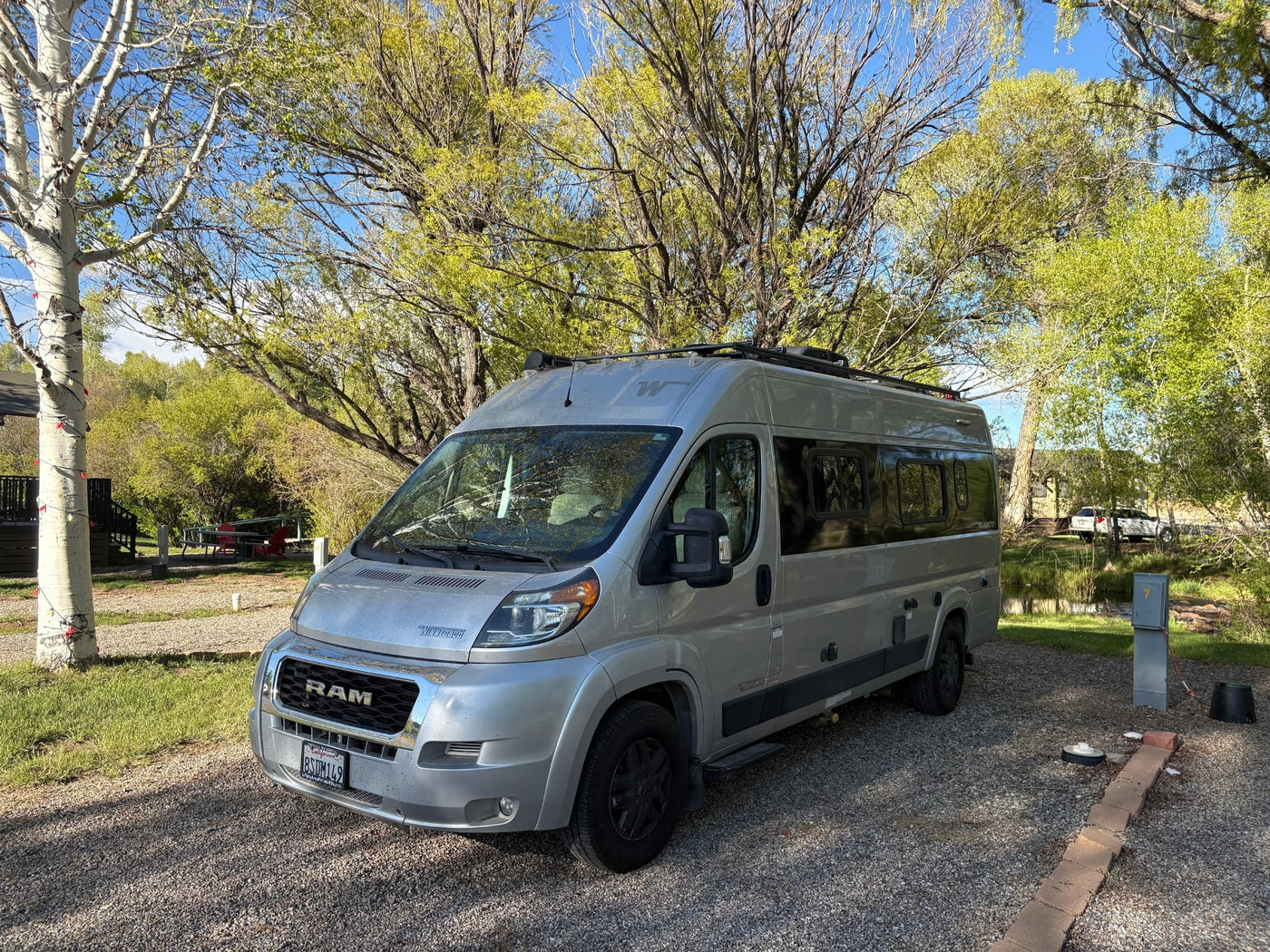 An RV rental, a silver Winnebago Travato, parked on a gravel campsite with lush trees and a blue sky overhead. — photo 4