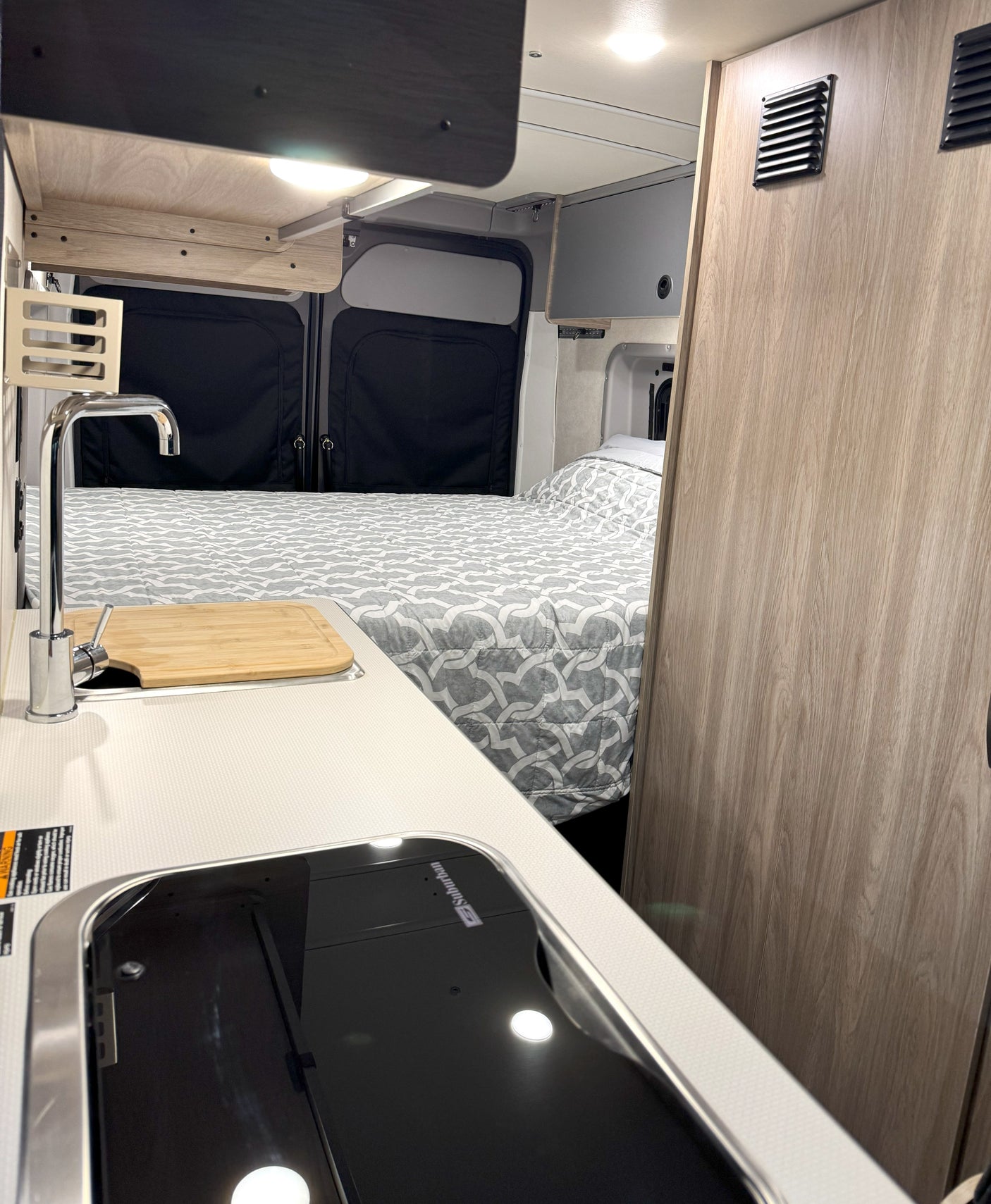 Interior of an rv rental: compact kitchen with cooktop, sink, and wooden cutting board. Beyond is a bed with gray patterned comforter.