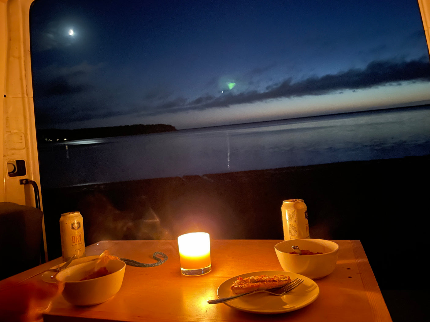 Cozy RV rental dinner by the water at night. Candlelight illuminates food, drinks, and a serene moonlit lake with a faint sky glow.