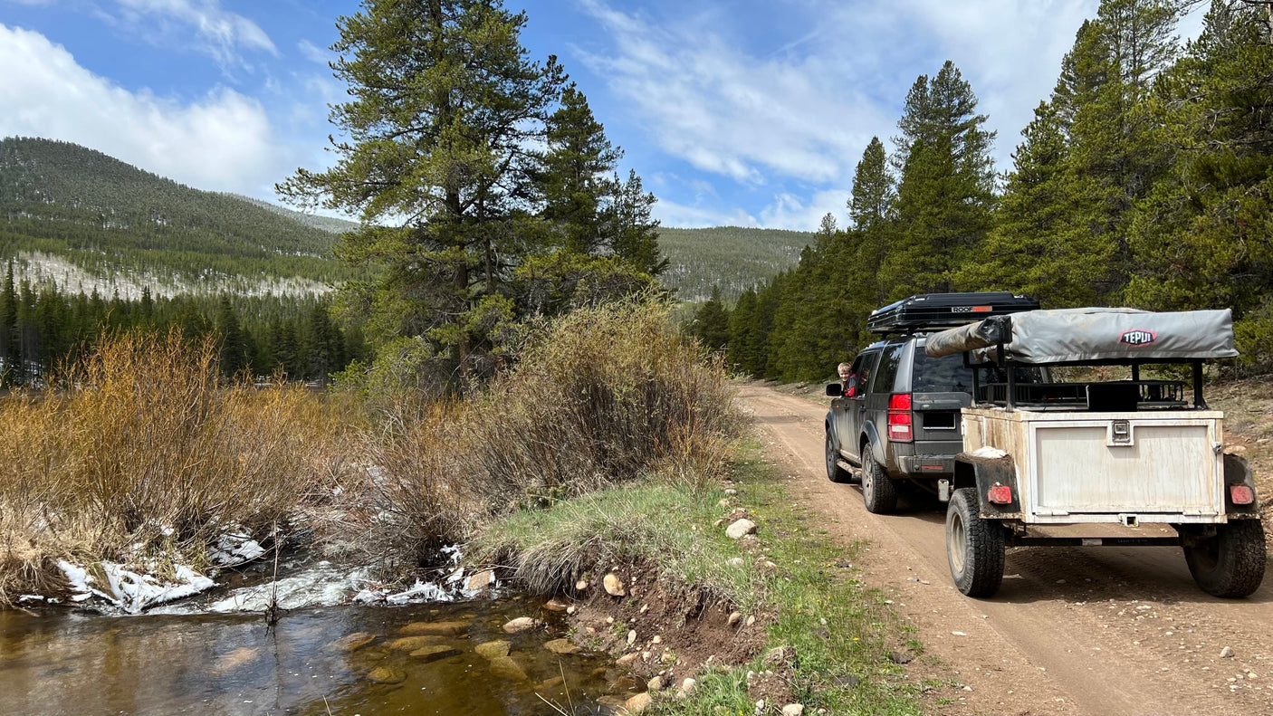 Overlanding adventure: SUV with roof-top tents & trailer, like an RV rental, on a dirt road next to a stream. Child visible.