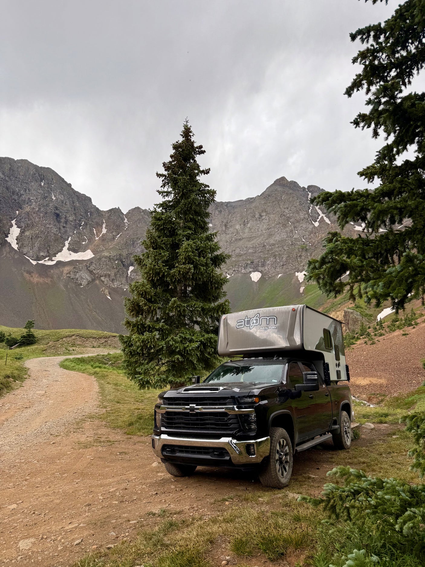 Black pickup with an 'atom' truck camper (RV rental) on a mountain dirt road, surrounded by pines and snowy peaks.