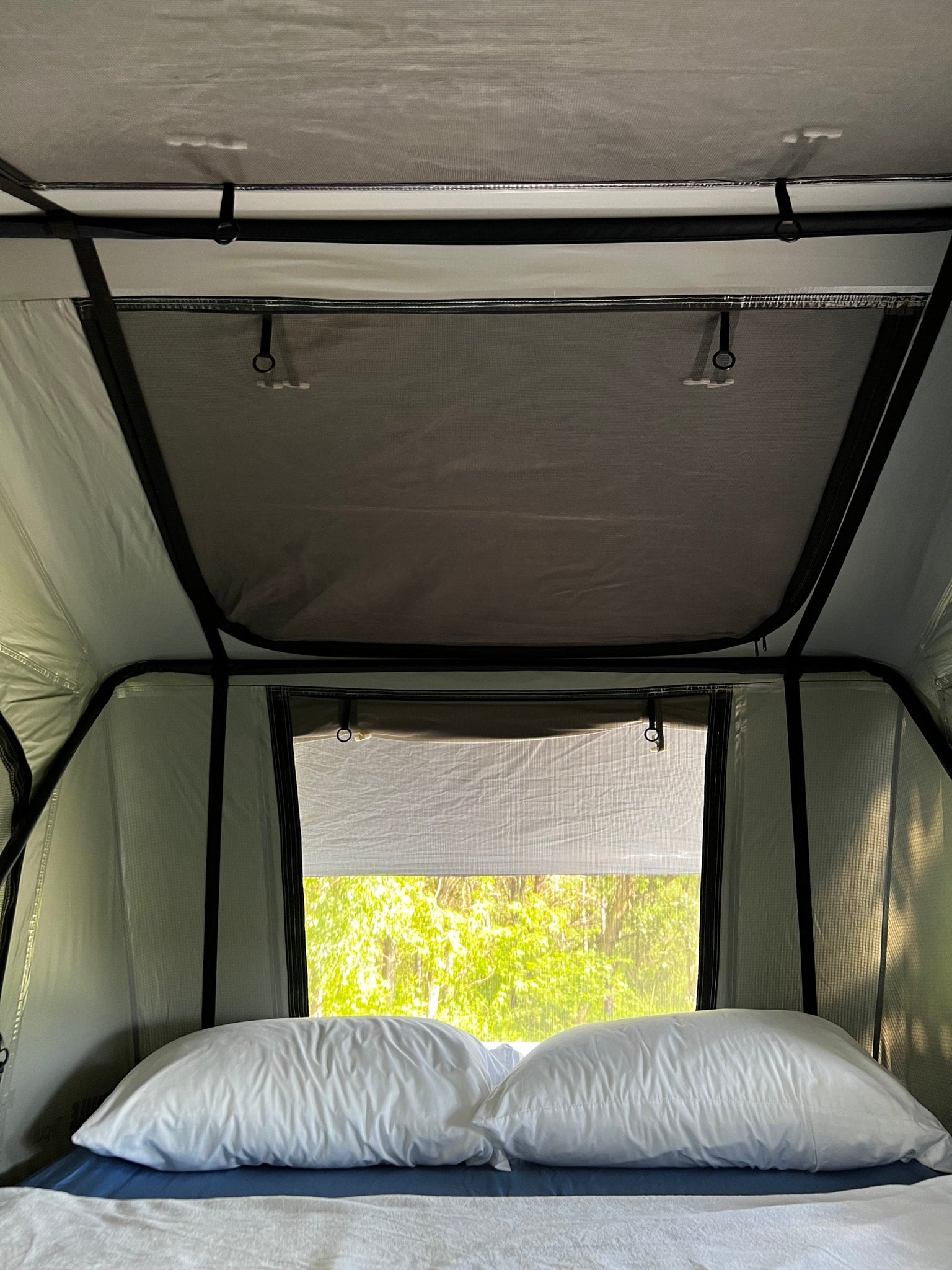 Inside a comfy RV rental: pillows on bed, bright nature view through mesh window in a rooftop tent/camper.