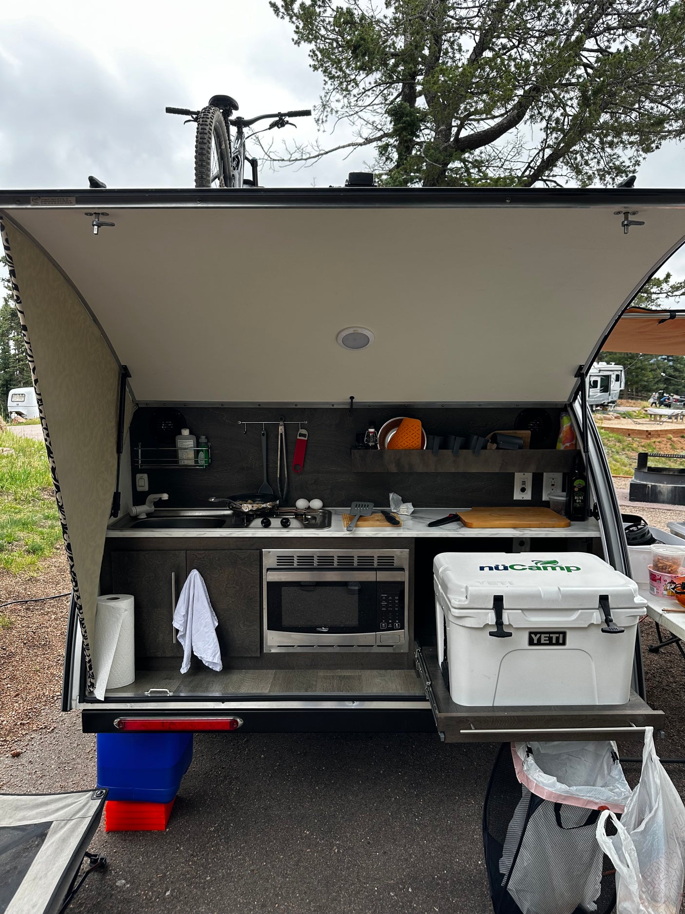 Enjoy the open outdoor kitchen of this rv rental camper at a campsite. Features a stove, microwave, and cooler, plus a bike on top.