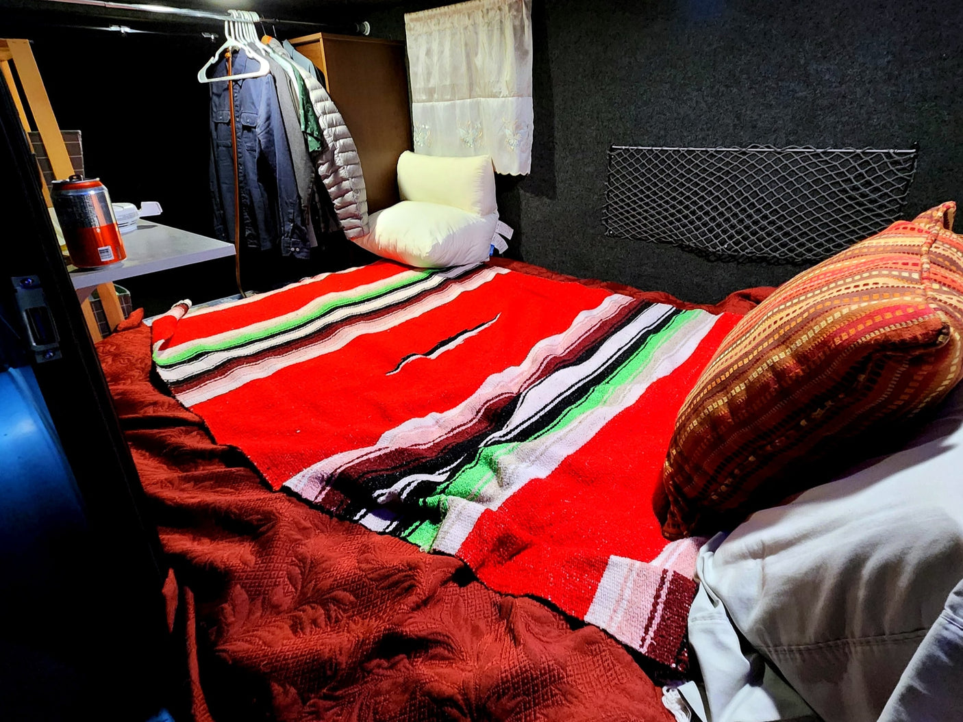 This RV rental features a cozy sleeping area with a vibrant red blanket, storage nets, and hanging clothes.