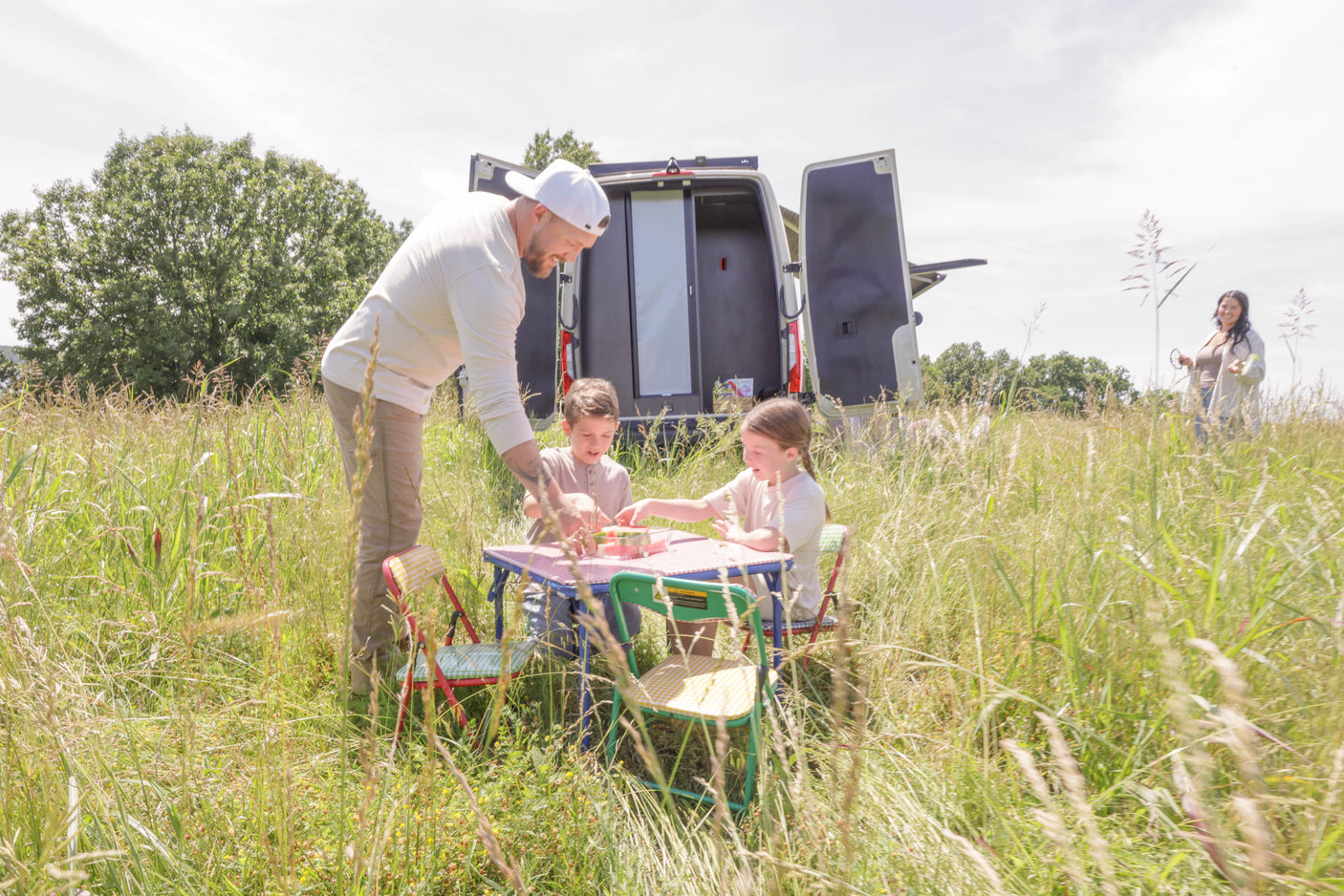 Family picnic with an RV rental: a man helps two children at a table in a grassy field, a woman stands by the open van.