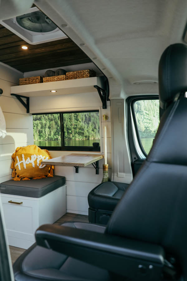Cozy RV rental interior: white walls, wood ceiling, shelf, window with lake view, fold-down table, and comfy seating.