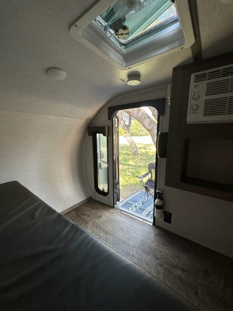 Inside an RV rental: compact living with a window view of trees, AC, and a cozy bed.
