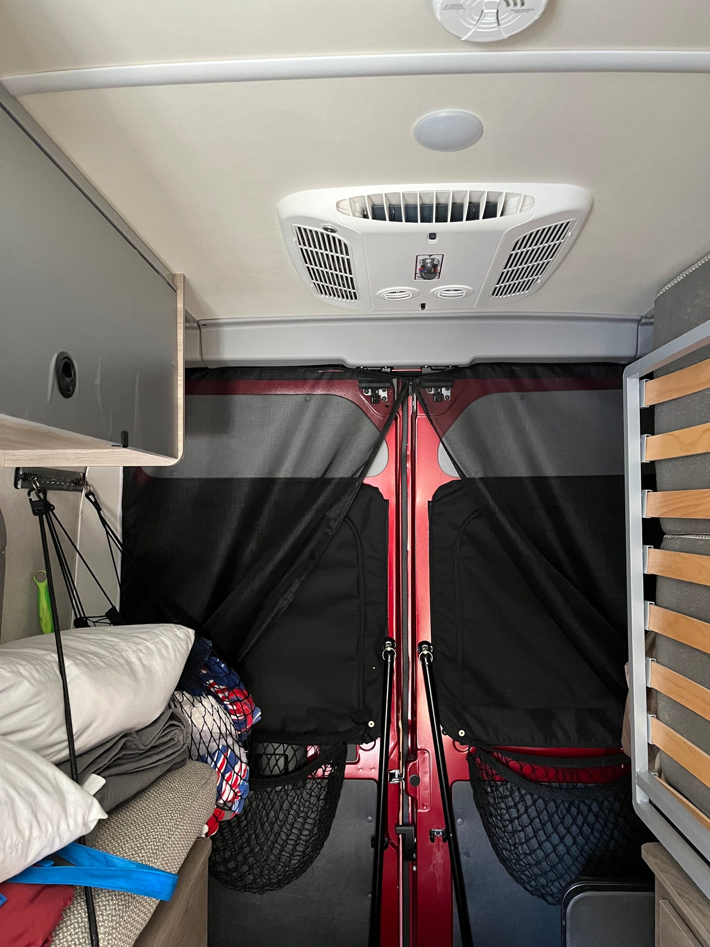 RV rental interior: Overhead AC unit, storage, stacked bedding, and fabric screens covering the red rear doors.