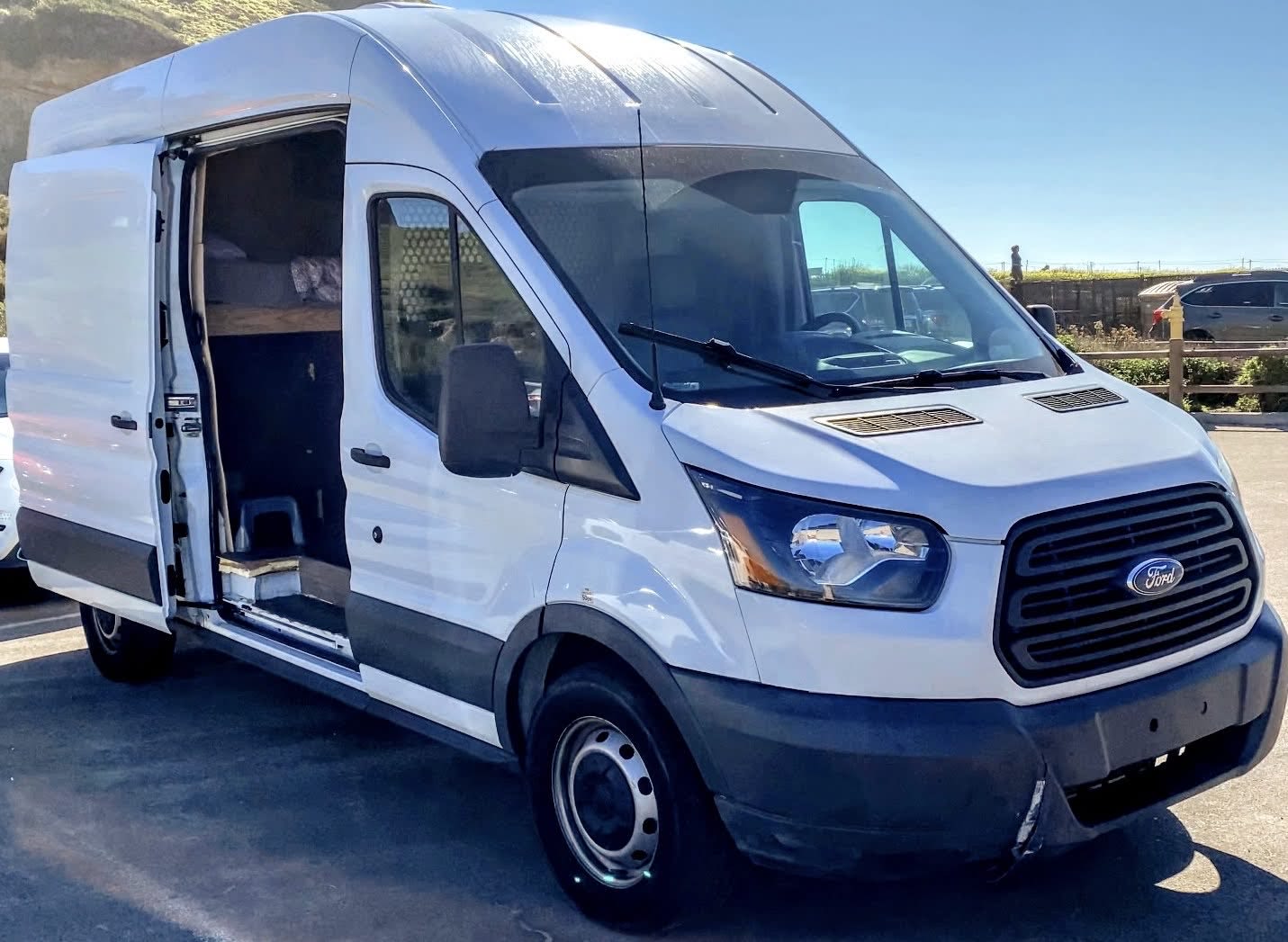 White Ford Transit camper van for RV rental, with its side door open revealing a cozy bed inside, perfect for adventure.