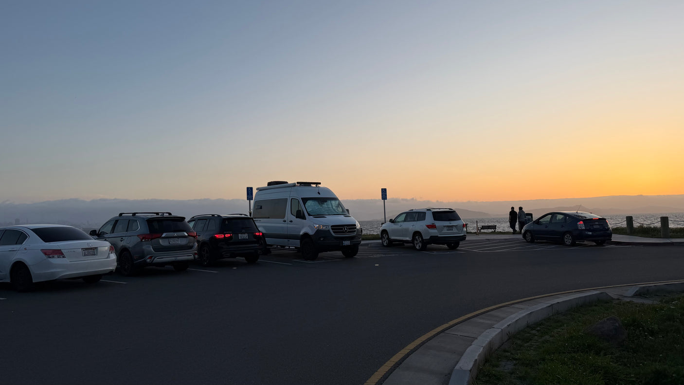 An RV rental, other vehicles, and people in a coastal parking lot at sunset, offering beautiful ocean views.