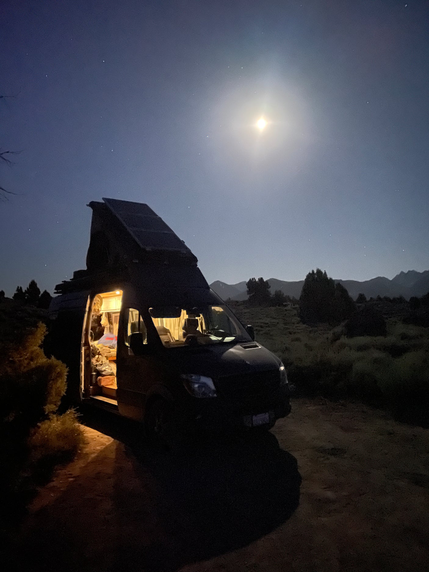 Nighttime serenity: an RV rental with its door open, glowing under the bright moon and stars, parked in a natural landscape.