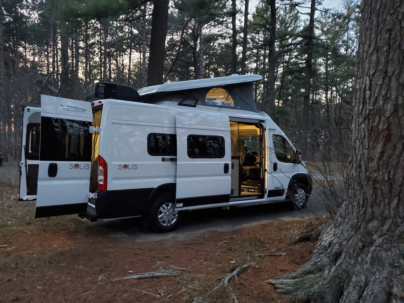 Experience nature with this Winnebago Solis rv rental parked in a forest. Doors open, pop-top up, ready for adventure.