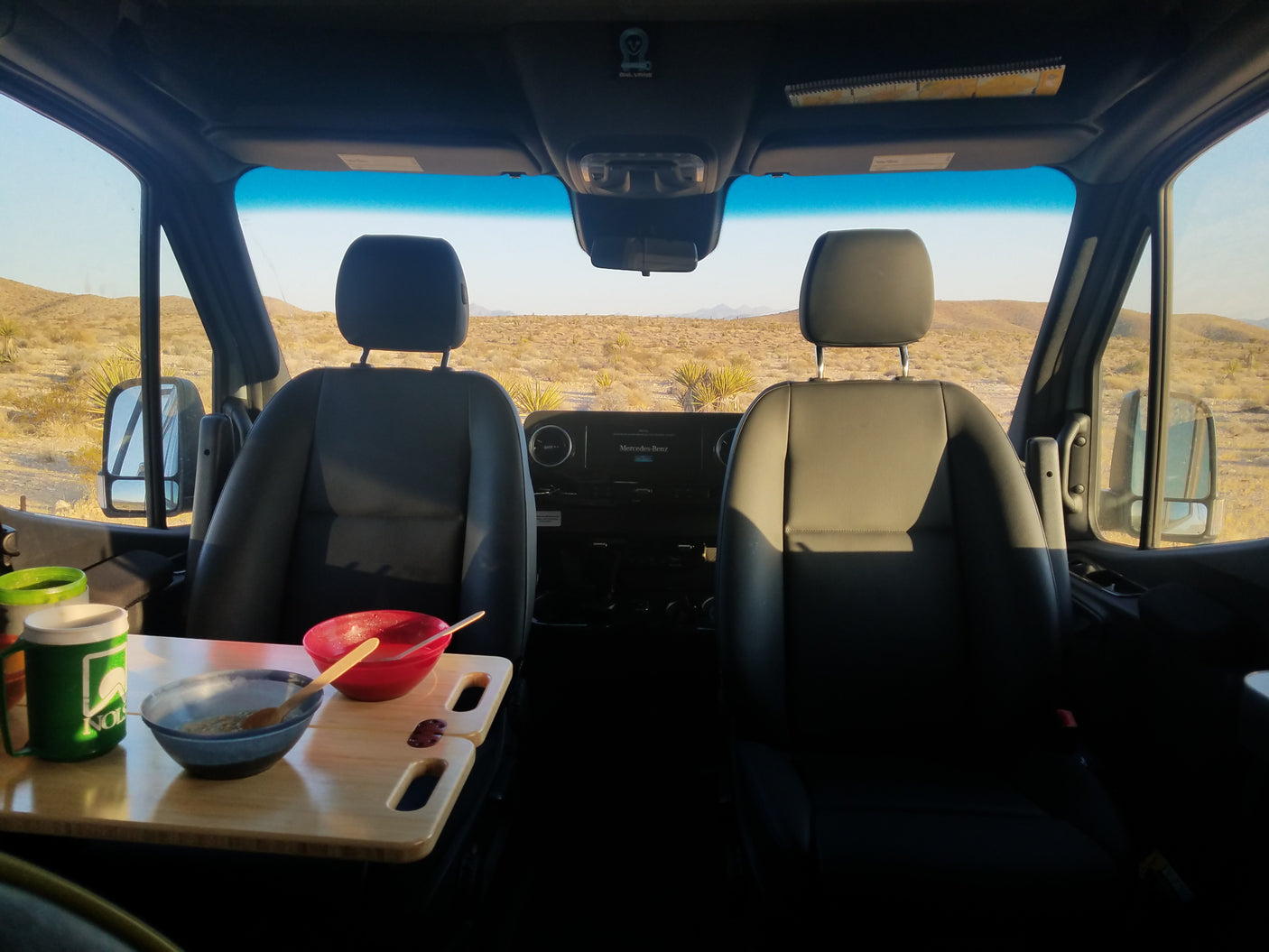 Inside an **rv rental** parked in the desert, two bowls and a mug sit on a table, with open views of sandy terrain and mountains.