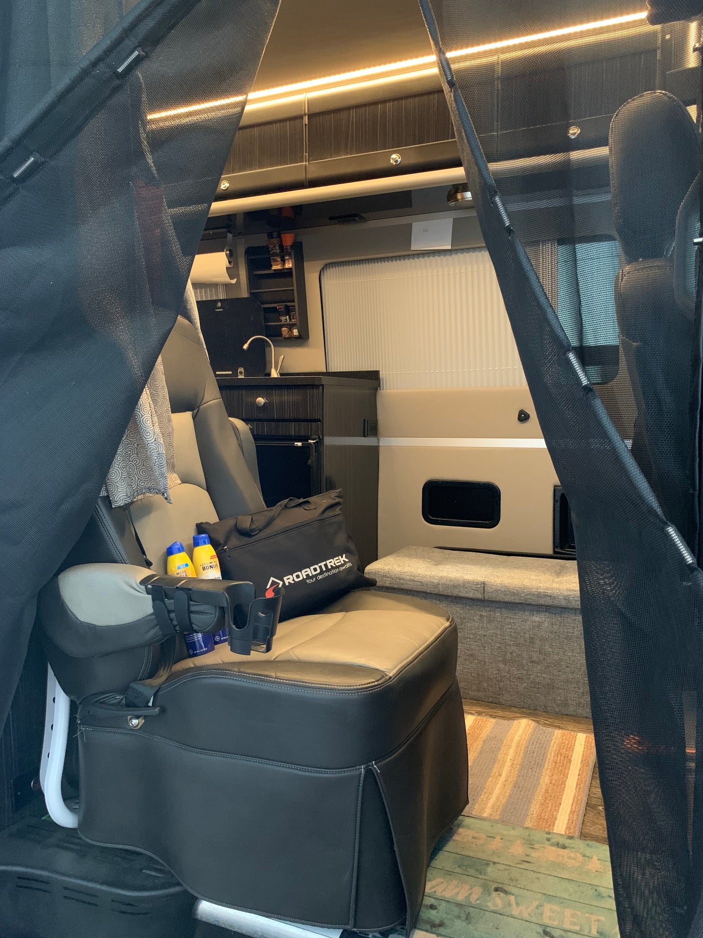 Inside an RV rental: a plush seat with a 'Roadtrek' bag and cupholders, a compact kitchen, and black mesh privacy screens.