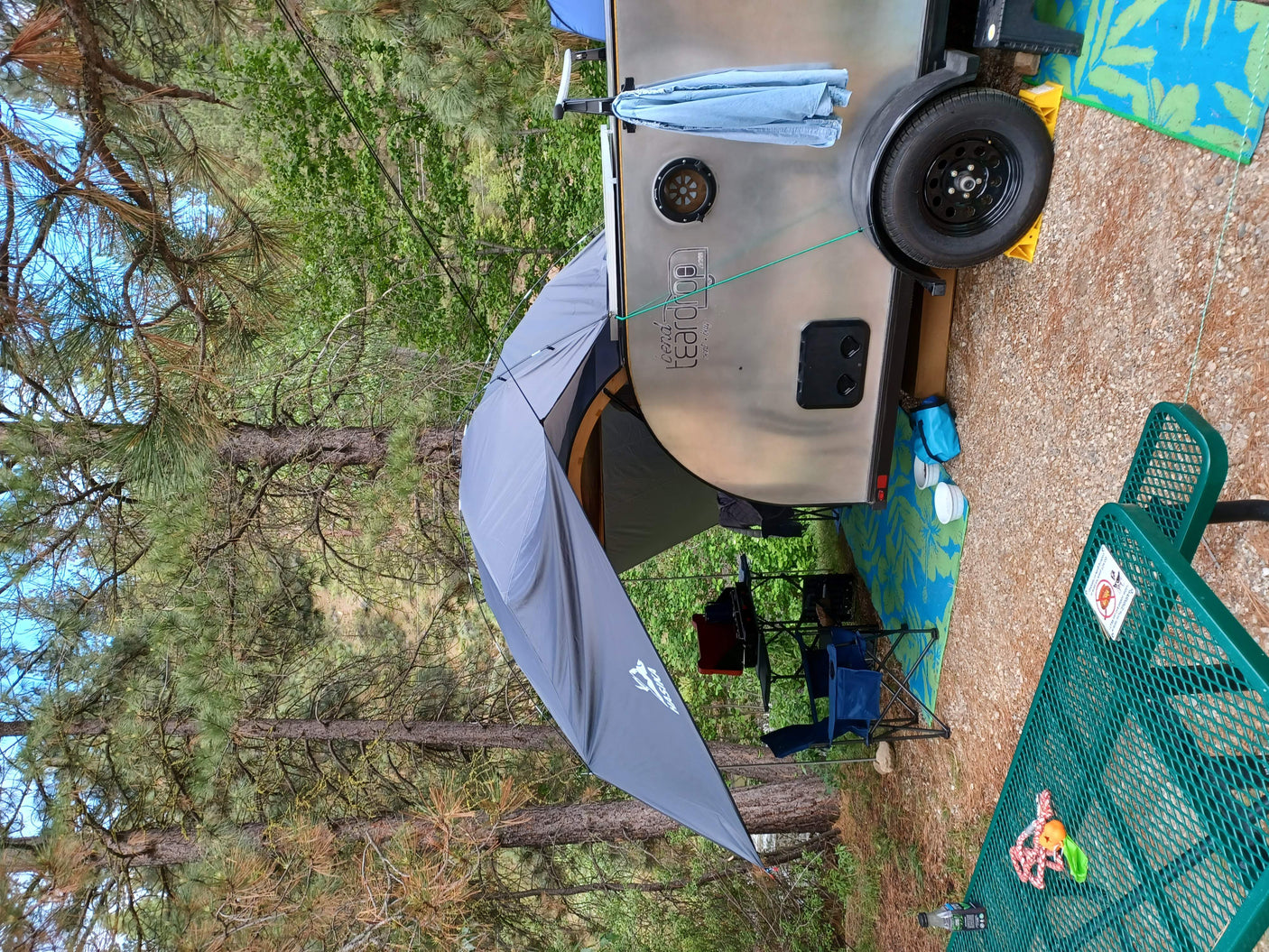 Teardrop RV rental set up with an attached awning, camping chairs, and a picnic table on gravel, nestled among pine trees.