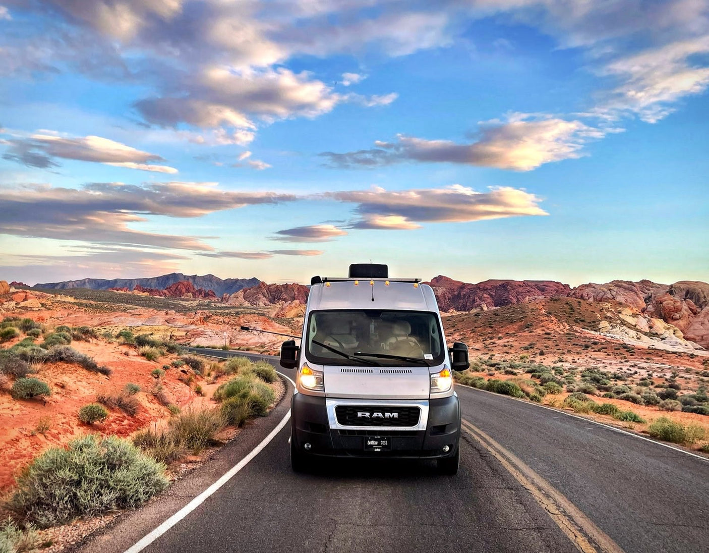 Scenic desert adventure: Silver RV rental on a road with stunning red mountains & a vibrant sky under scattered clouds.|Smiling woman on a sandy beach next to her RV rental van