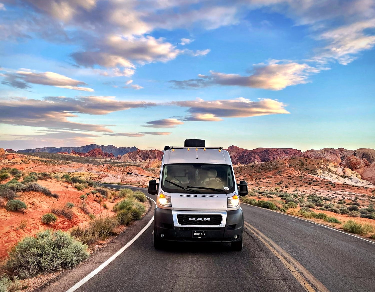 Scenic desert adventure: Silver RV rental on a road with stunning red mountains & a vibrant sky under scattered clouds.|Smiling woman on a sandy beach next to her RV rental van