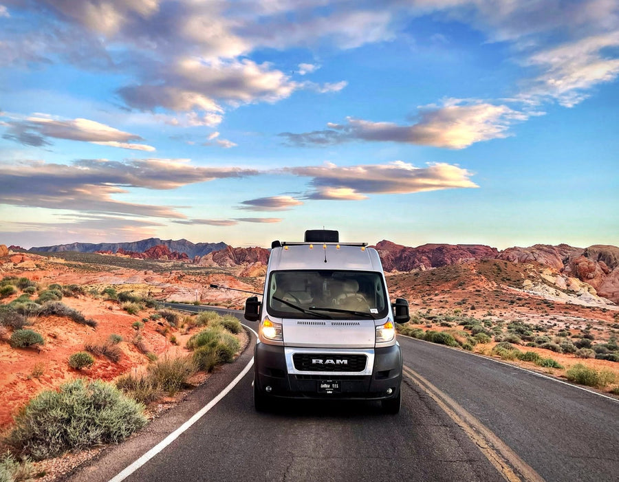 Scenic desert adventure: Silver RV rental on a road with stunning red mountains & a vibrant sky under scattered clouds.|Smiling woman on a sandy beach next to her RV rental van