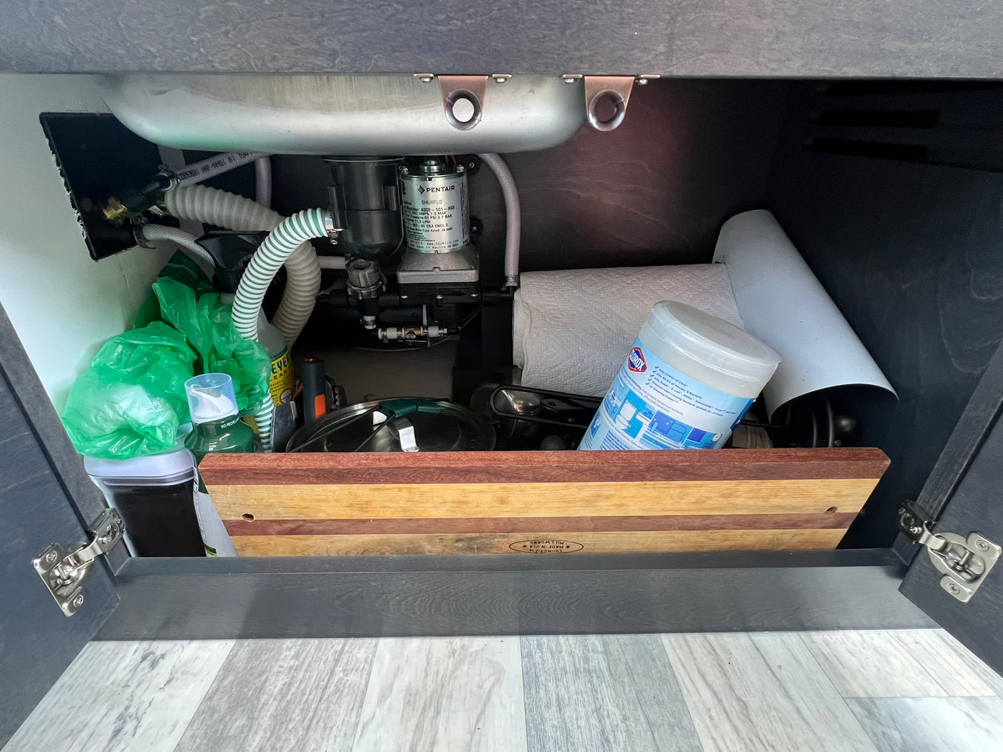 RV rental kitchen cabinet: compact sink, Shurflo water pump, plumbing, and stored essentials like paper towels & cookware.