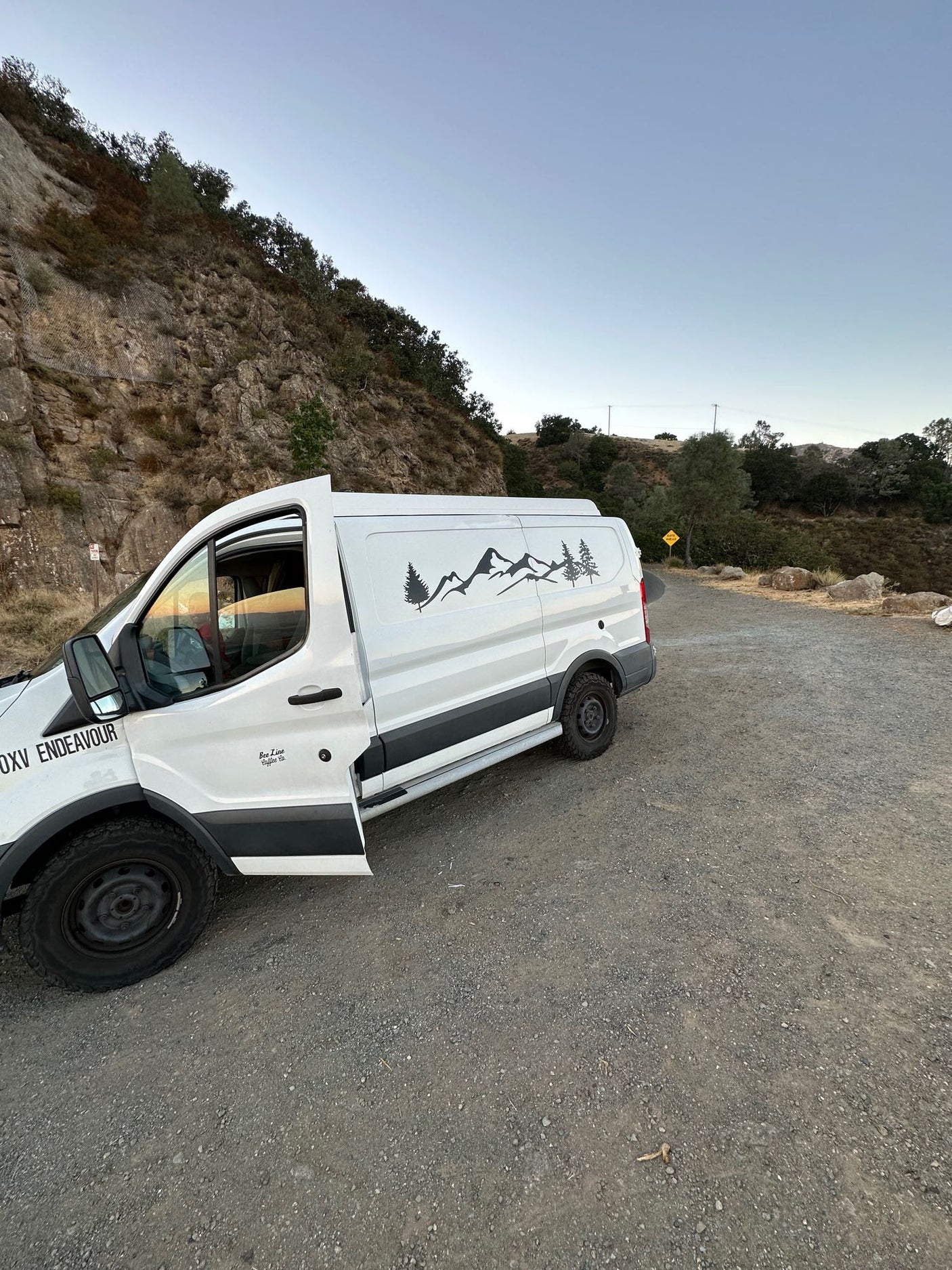 White camper van (RV rental) with mountain decals parked on a gravel road beside a rocky hillside.