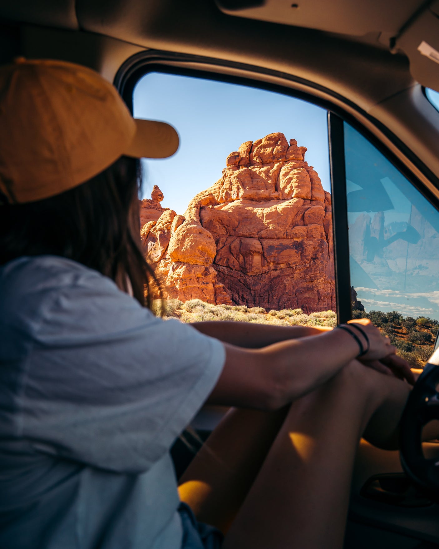 From an RV rental, a traveler with a yellow cap admires the vast red rock desert landscape through the vehicle window.