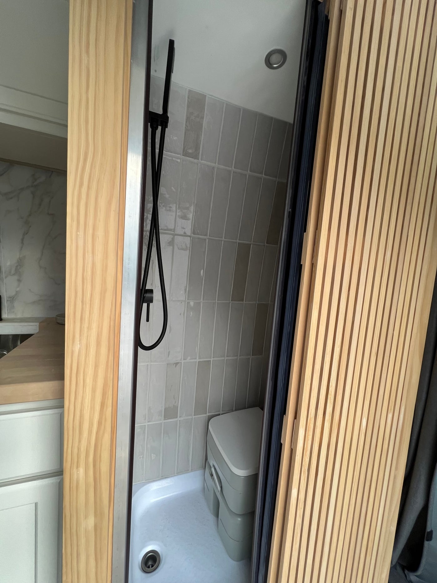 An RV rental features a compact wet bath with black shower, grey tiled walls, and a portable composting toilet. — photo 8