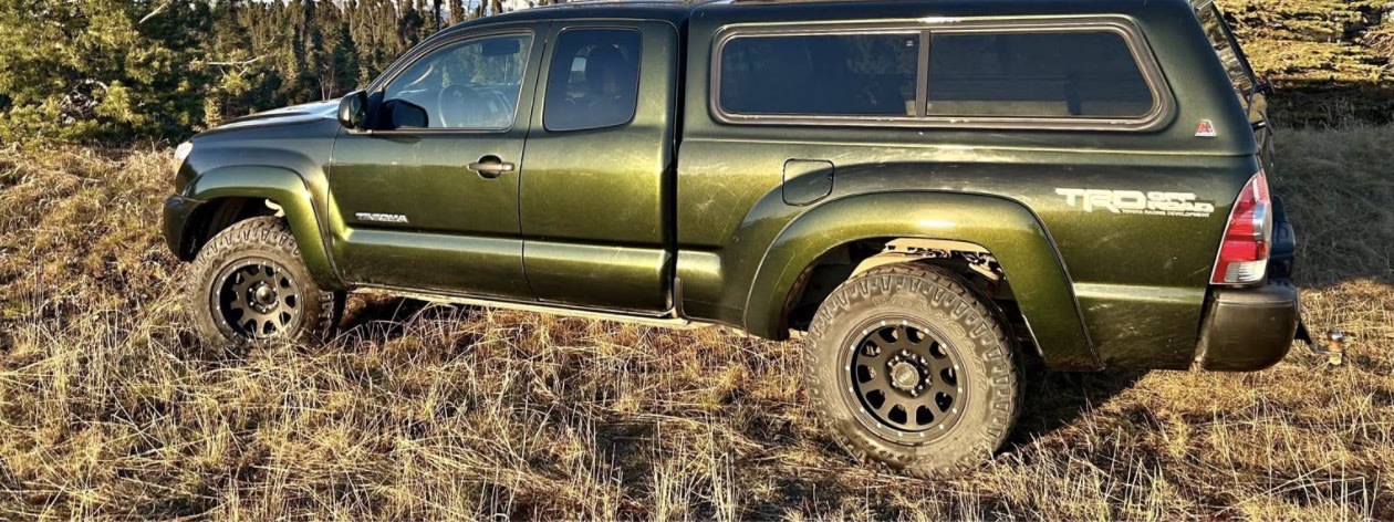 Olive green Toyota Tacoma pickup with camper shell off-road in dry grass. Great for an rv rental adventure!