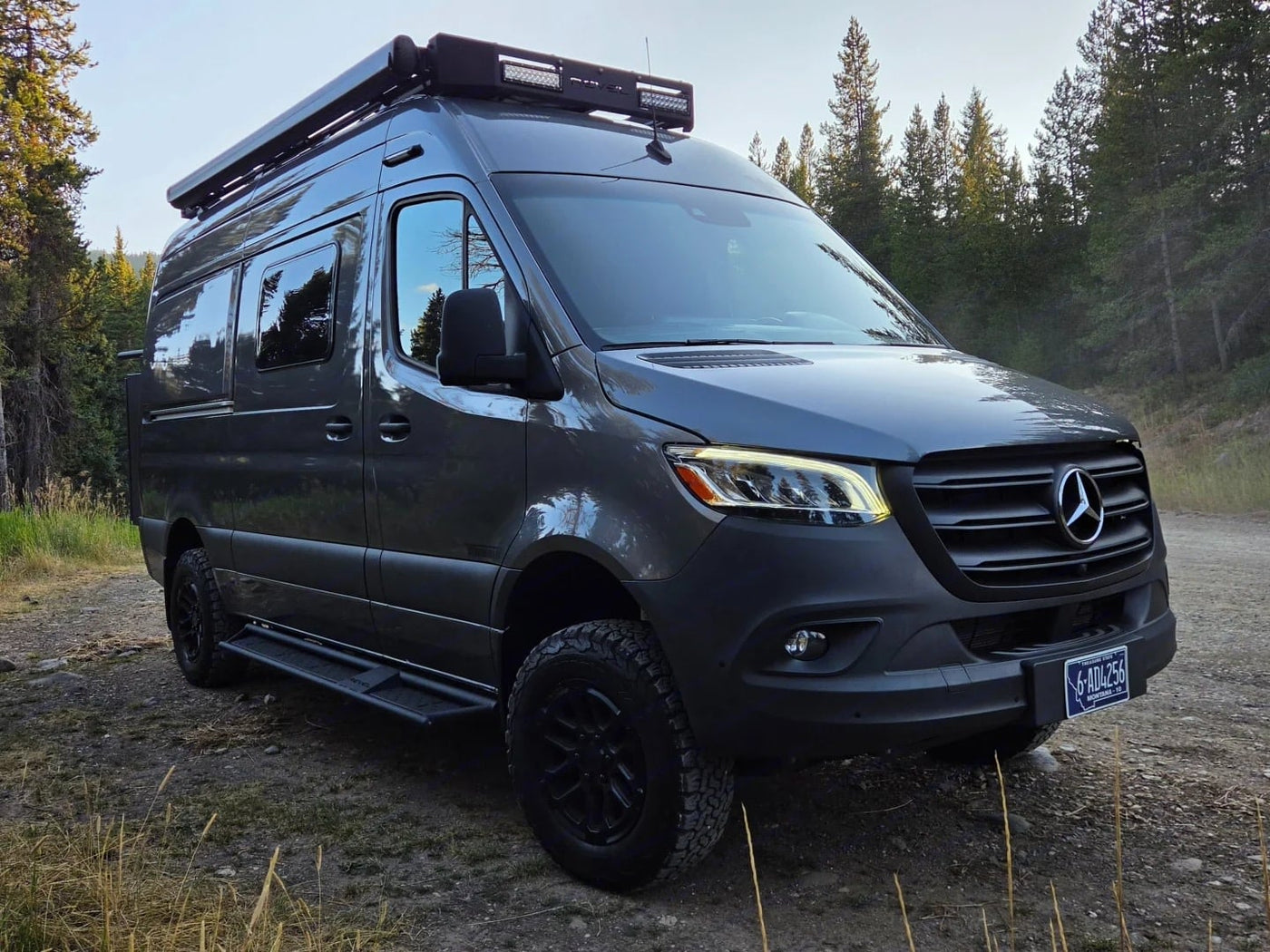 ready for off-road adventure.|Adventure-ready dark grey Mercedes Sprinter rv rental. This off-road camper van is parked on a dirt path by tall trees.|Black Mercedes Sprinter rv rental on a Montana lake shore — photo 3
