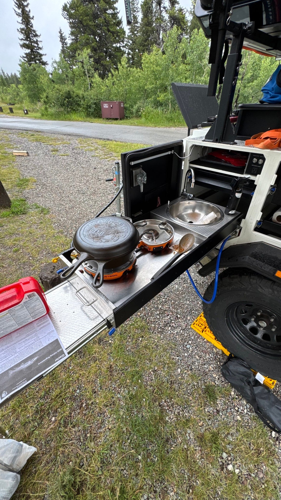 This RV rental's outdoor kitchen features a stove, cast iron pan, and sink, set up in a wooded campground.