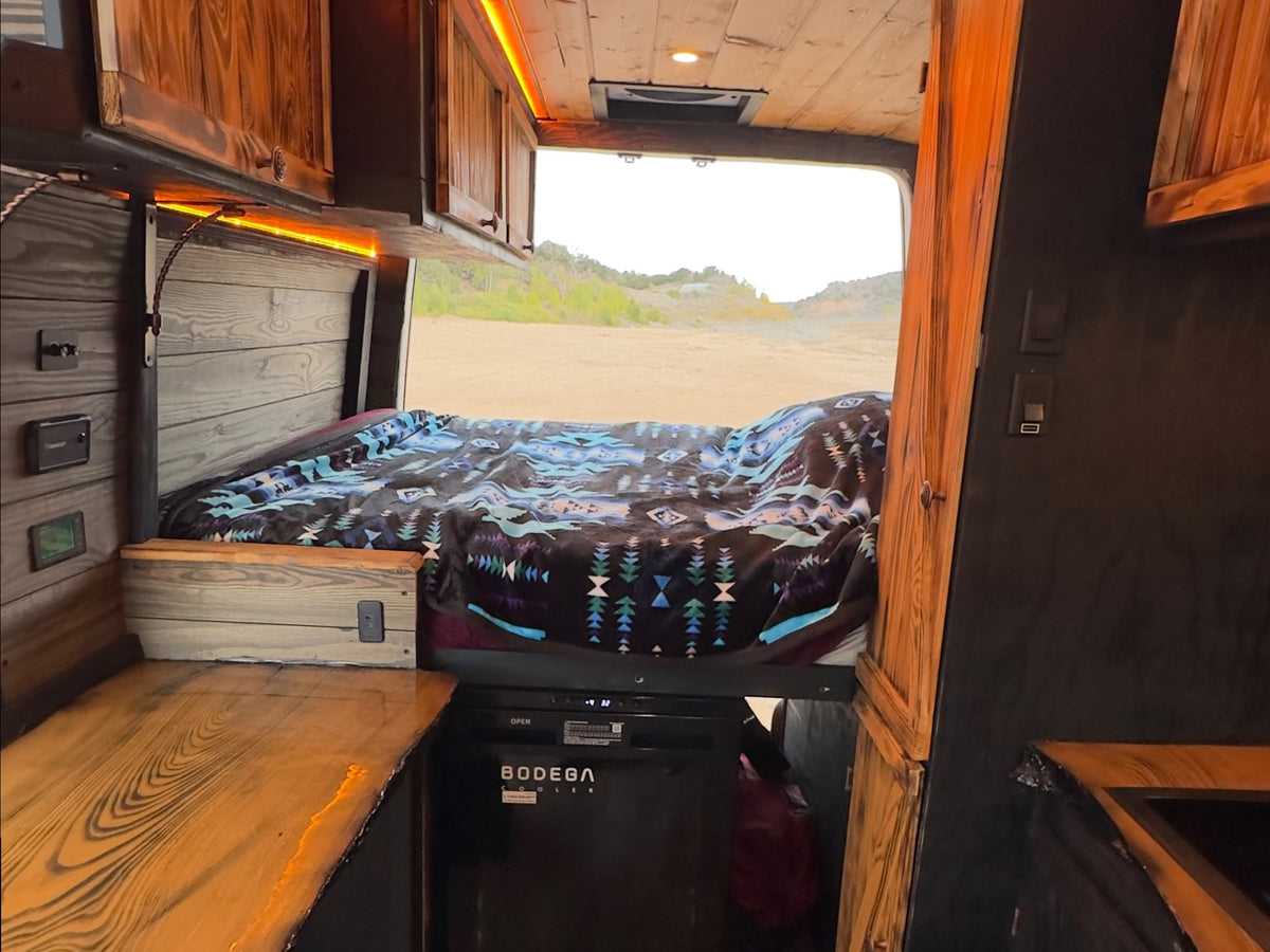 parked on dirt against a backdrop of green hills and a cloudy sky.|Cozy RV rental interior with a patterned blanket on the bed — photo 6