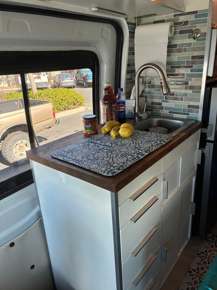Compact kitchen in an rv rental camper van, with a sink, wooden counter, fruit, and a patterned mat.