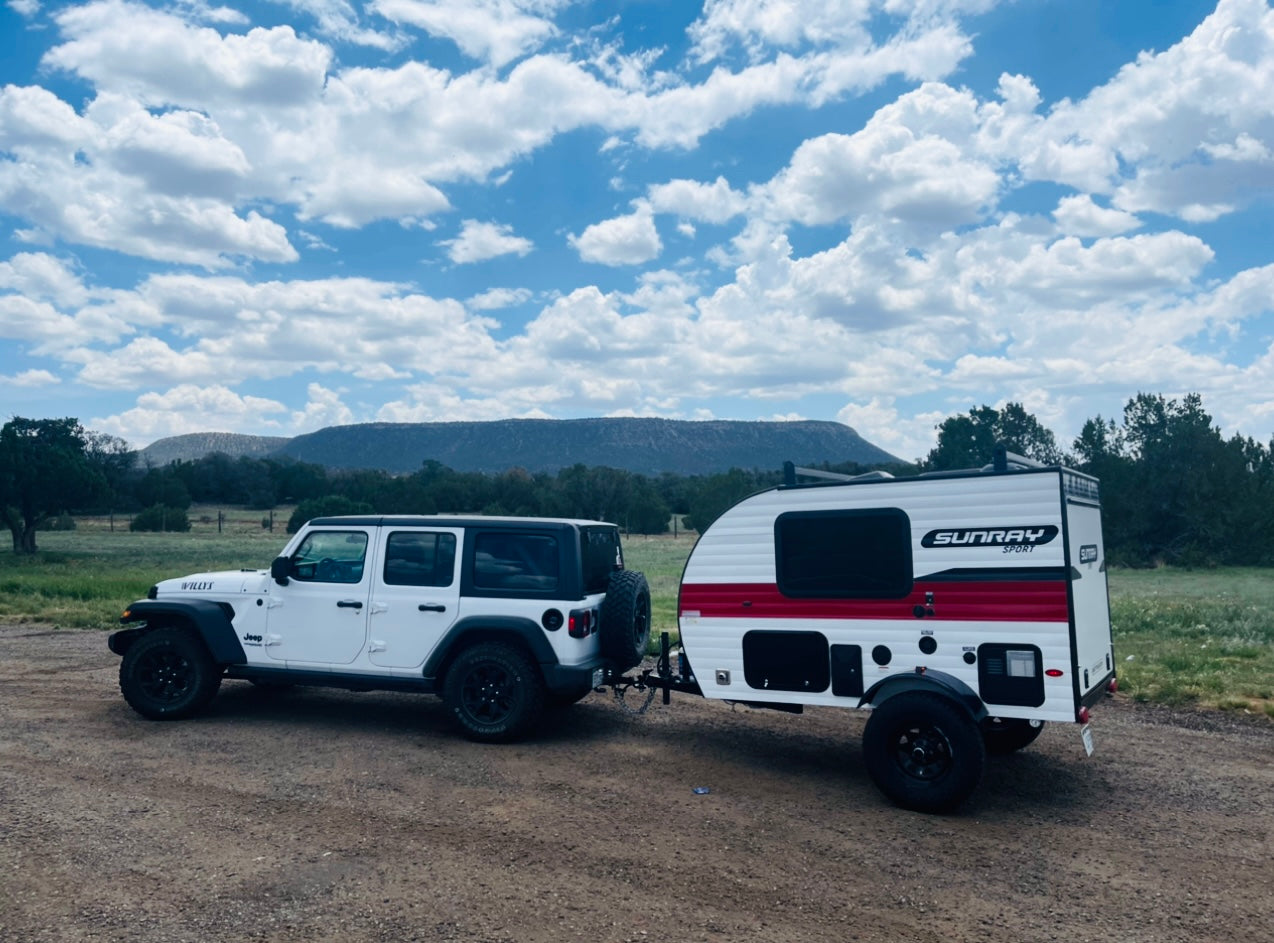 Adventure-ready rv rental: White Jeep Willys towing a Sunray Sport camper on a dirt road, mountains & clouds.