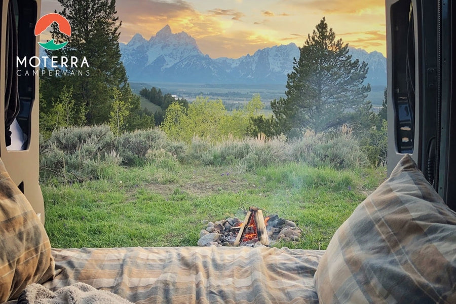 Cozy RV rental view: Inside a camper van, enjoy snow-capped mountains, a sunset sky, and a crackling campfire.