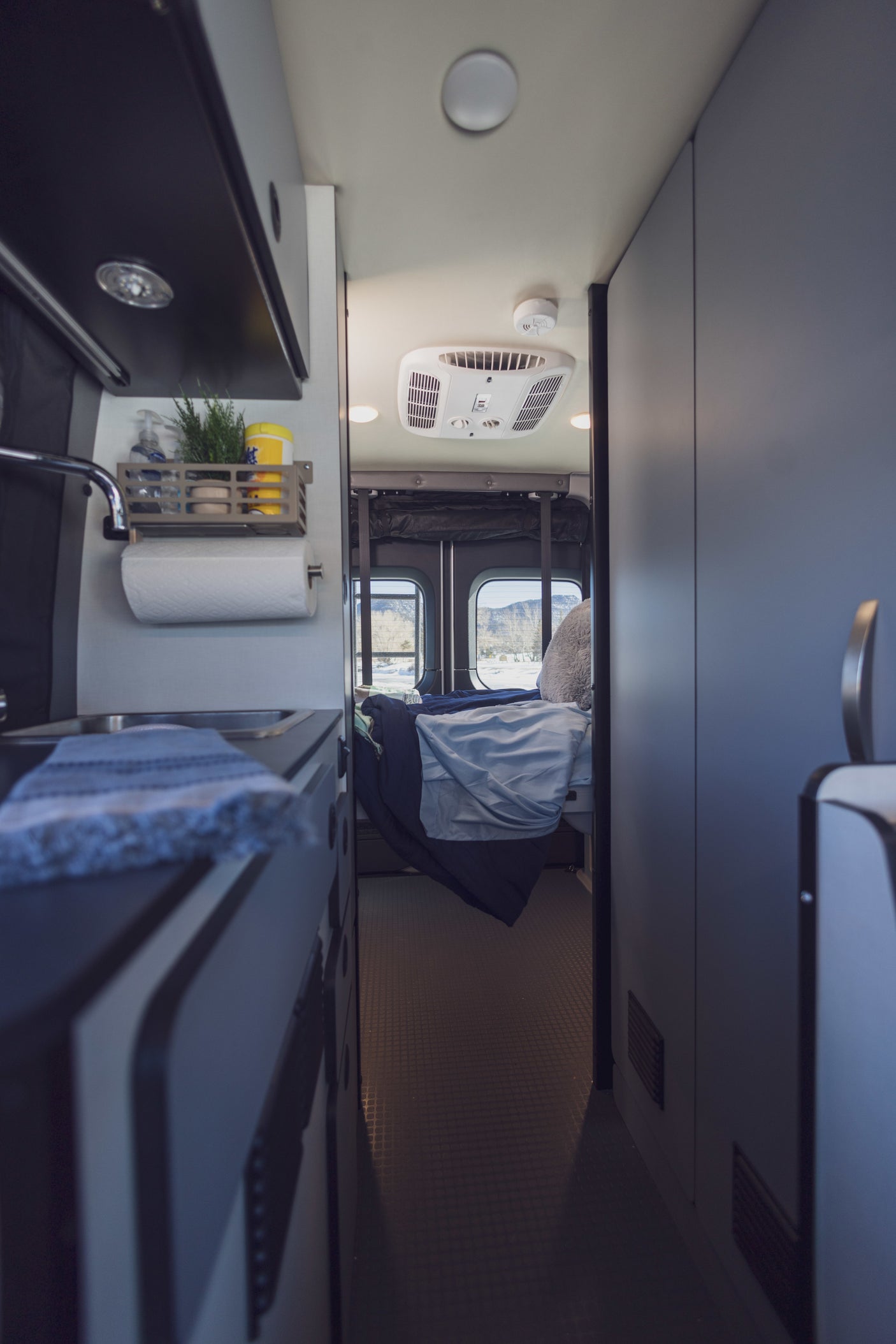 This RV rental interior showcases a compact kitchen area on the left and a cozy bedroom with rear window views, ideal for mobile living.