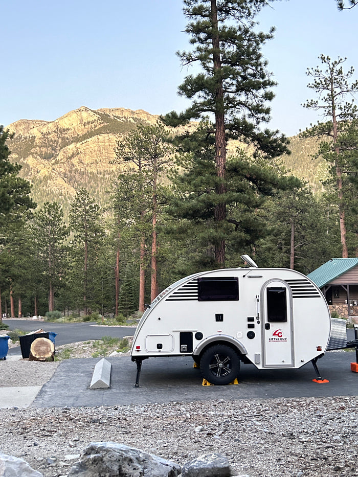 A white Little Guy rv rental teardrop trailer at a scenic mountain campsite, surrounded by towering pine trees.
