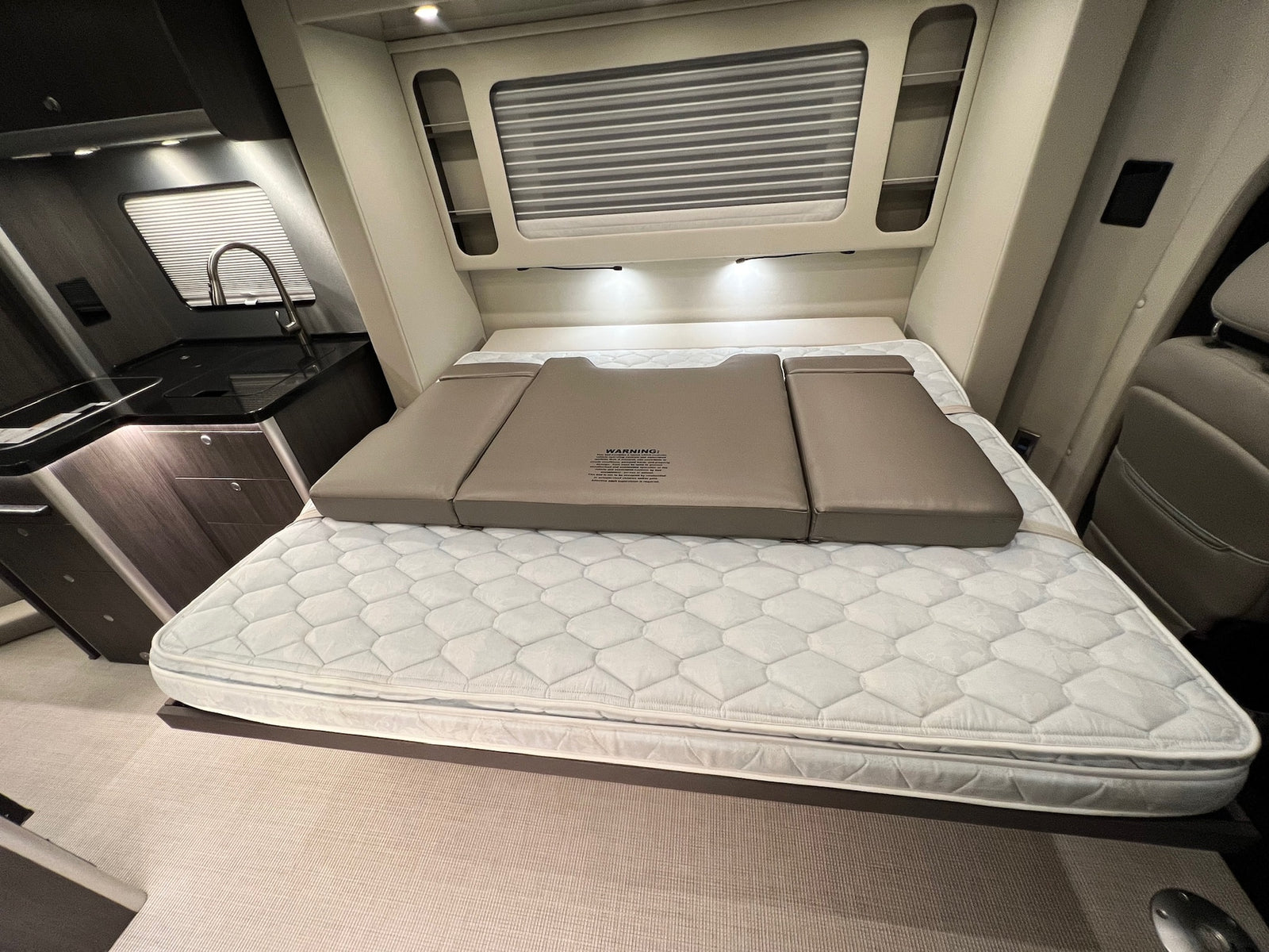 RV rental interior: A bed with a foldable extension, a compact kitchen with a sink on the left, and cabin seating on the right.