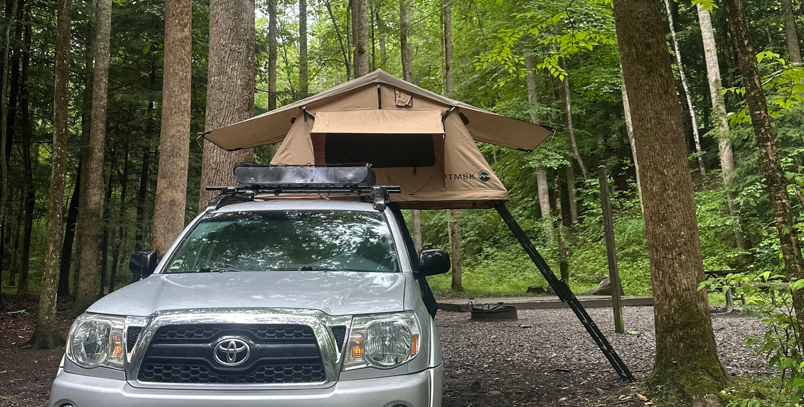Enjoy nature! This truck with a rooftop tent offers a unique alternative to an RV rental for camping in the woods.|A silver pickup truck with a TMBK rooftop tent set up for camping in a lush forest campsite