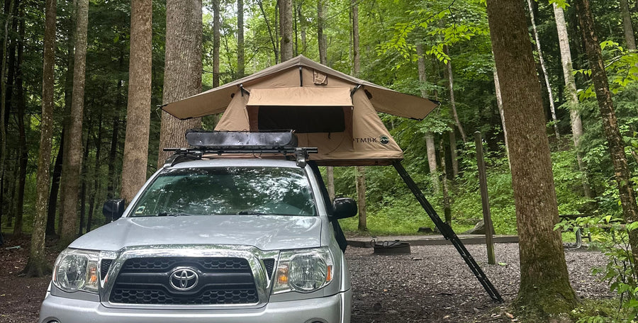 Enjoy nature! This truck with a rooftop tent offers a unique alternative to an RV rental for camping in the woods.|A silver pickup truck with a TMBK rooftop tent set up for camping in a lush forest campsite