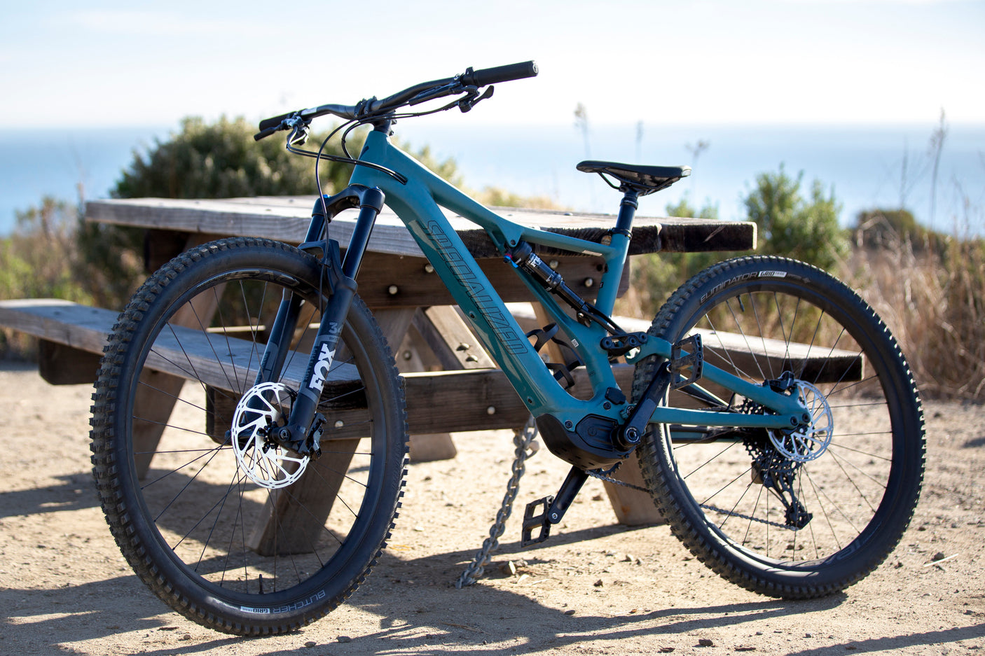 A teal Specialized e-mountain bike is parked by a picnic table, with an ocean view in the background. Perfect for RV rental adventures.