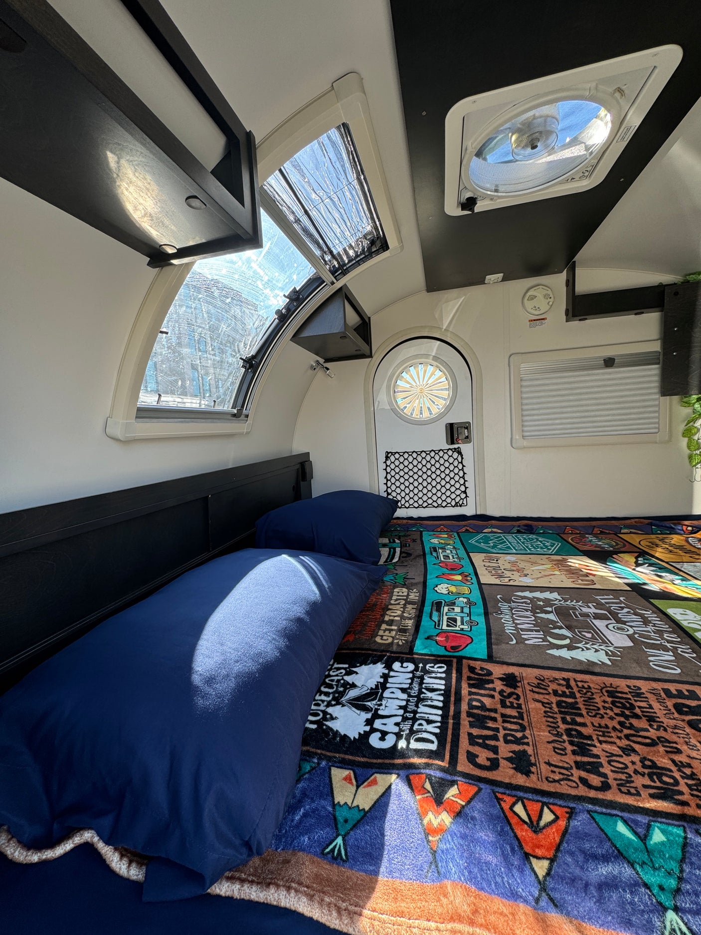 Inside a compact rv rental camper with a bed, "Camping Rules" blanket, bright windows, overhead storage, and a ceiling fan.