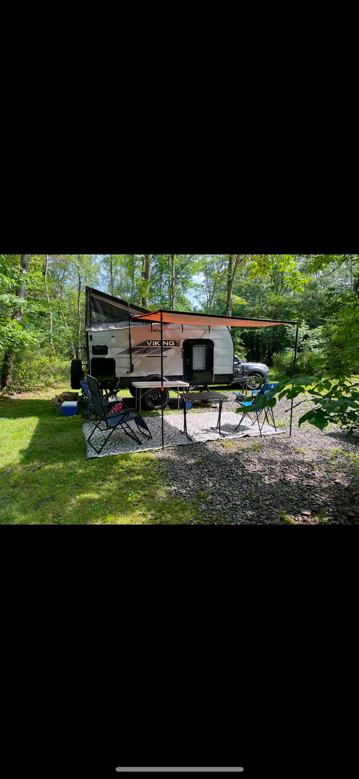 An RV rental, a Viking travel trailer, set up in a wooded campsite with an orange awning, chairs, and tables.