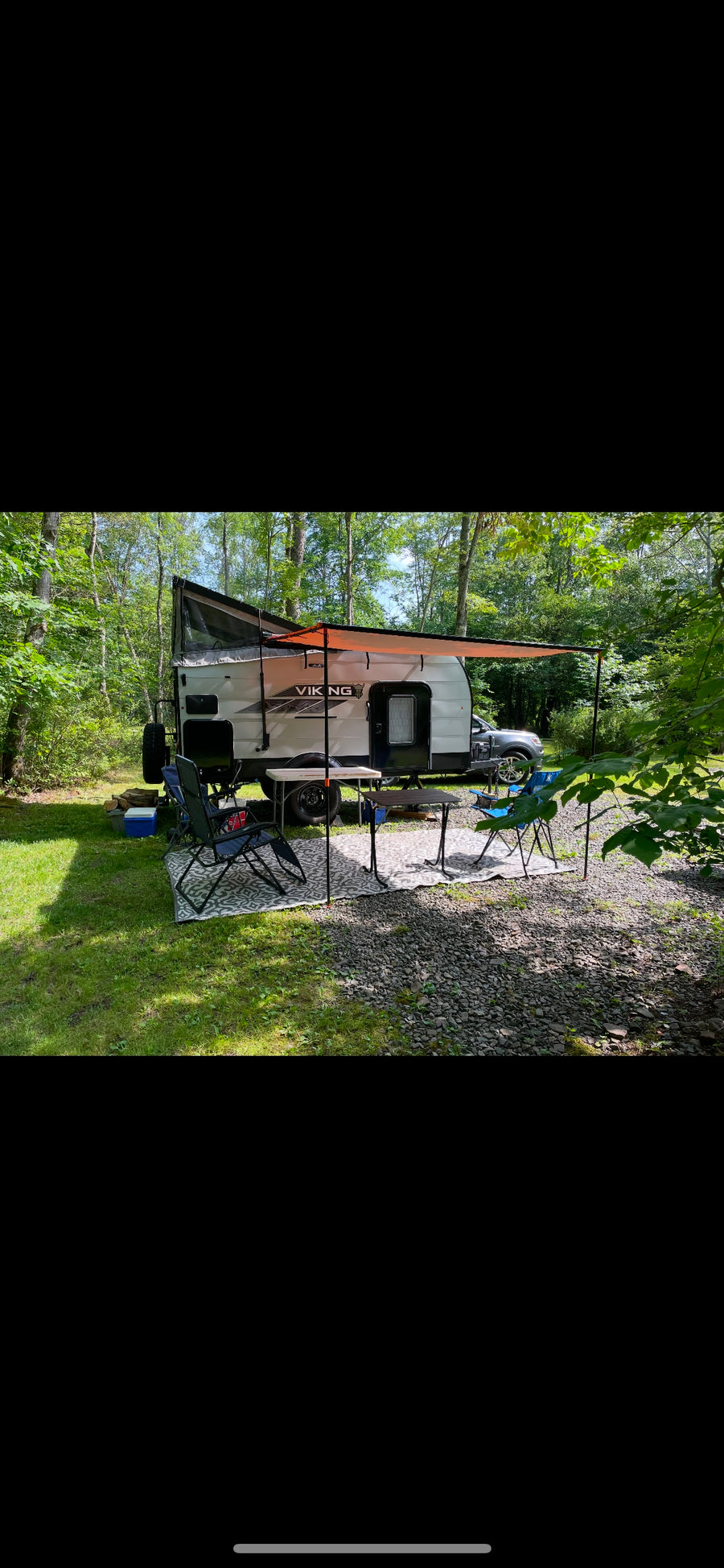 An RV rental, a Viking travel trailer, set up in a wooded campsite with an orange awning, chairs, and tables.