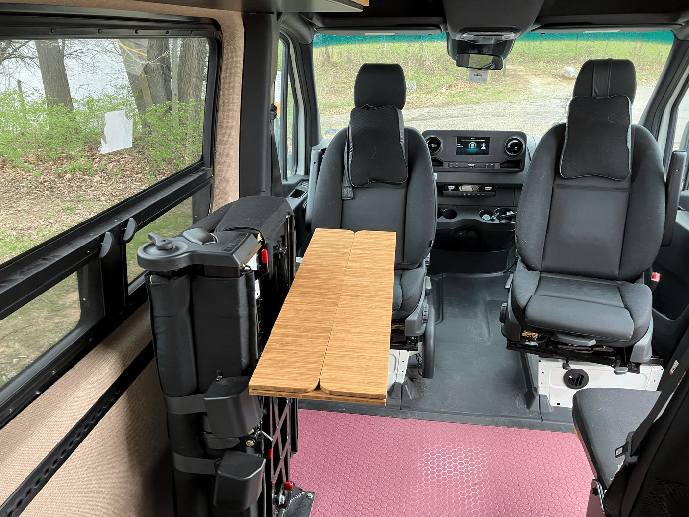Inside an RV rental, showing the front seats, a foldable table, and a mobility lift, with trees visible outside.