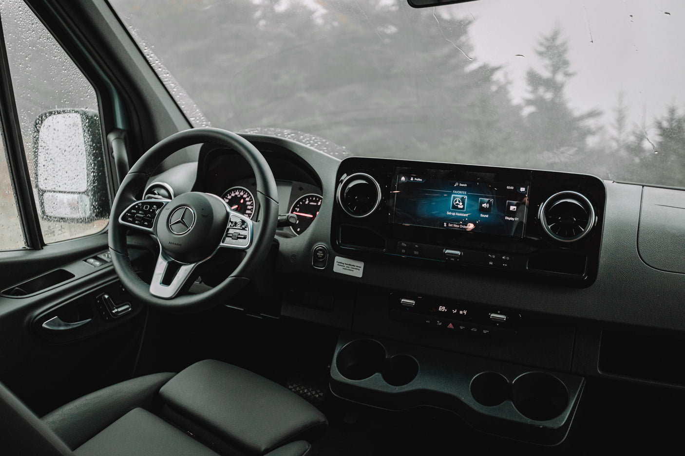 Interior of a Mercedes rv rental: dashboard, steering wheel, and infotainment system. Rain spatters the windshield, showing blurred trees.