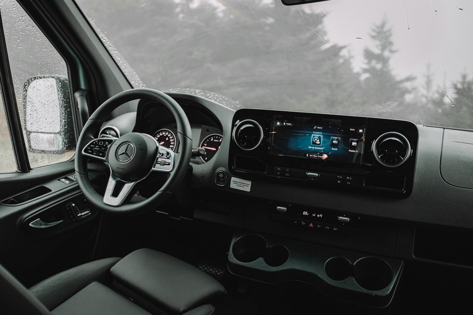 Interior of a Mercedes rv rental: dashboard, steering wheel, and infotainment system. Rain spatters the windshield, showing blurred trees.