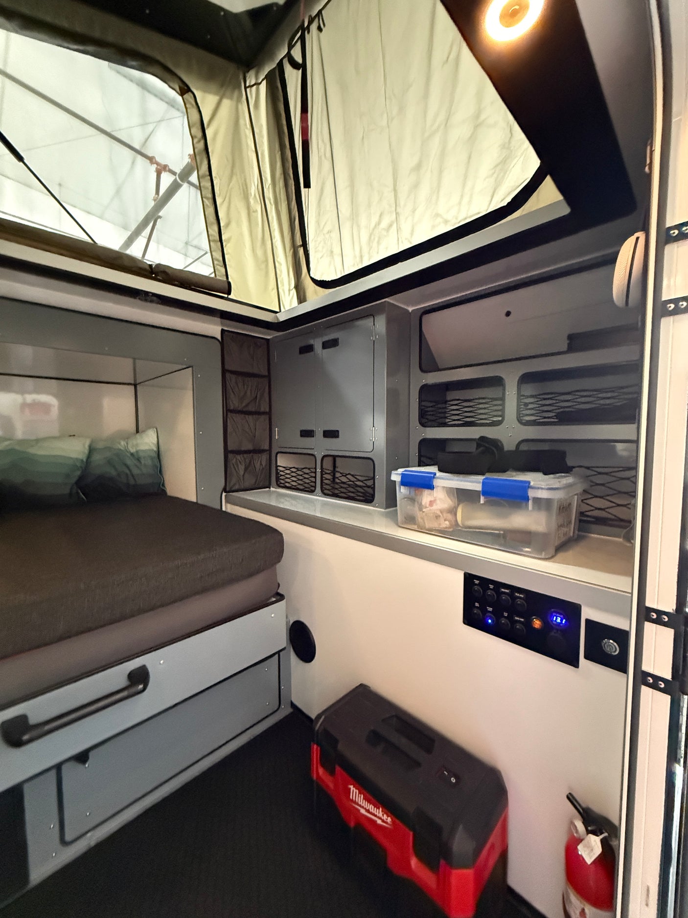 Interior of an RV rental pop-top camper, showing a bed, organized storage, an electrical panel, and a Milwaukee toolbox.