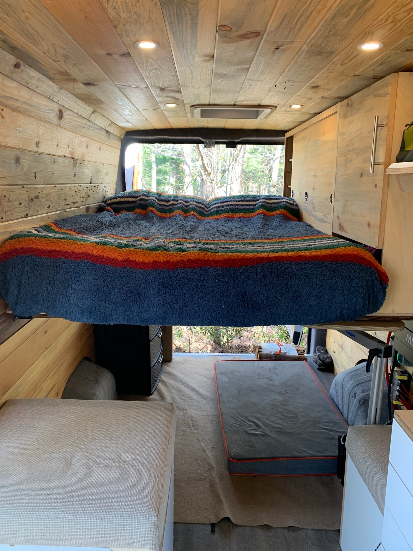 Cozy camper van interior for RV rental. Features a raised bed with striped blanket, wood paneling, and storage. Ideal for travel adventures.
