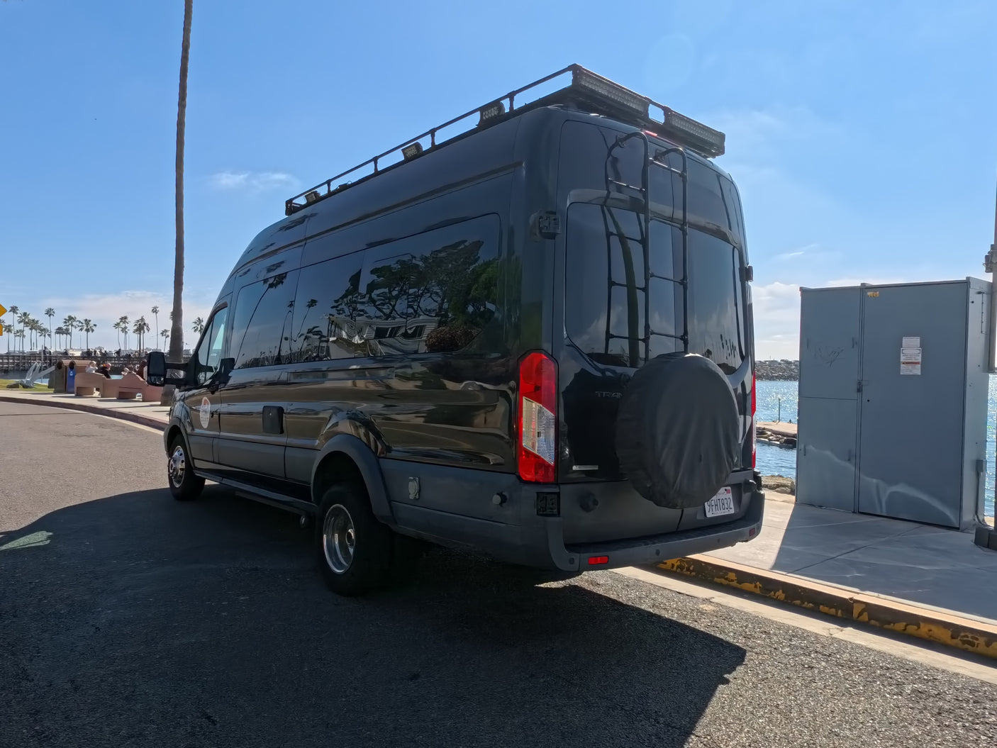 Sleek black rv rental conversion van with roof rack, ladder, and spare tire, parked by a sunny harbor.